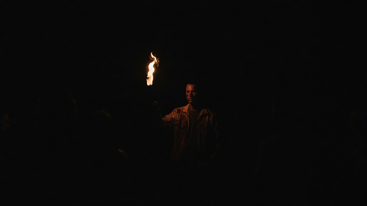 A lone performer in total darkness holds up a single lit torch, face barely warmed by the glow.