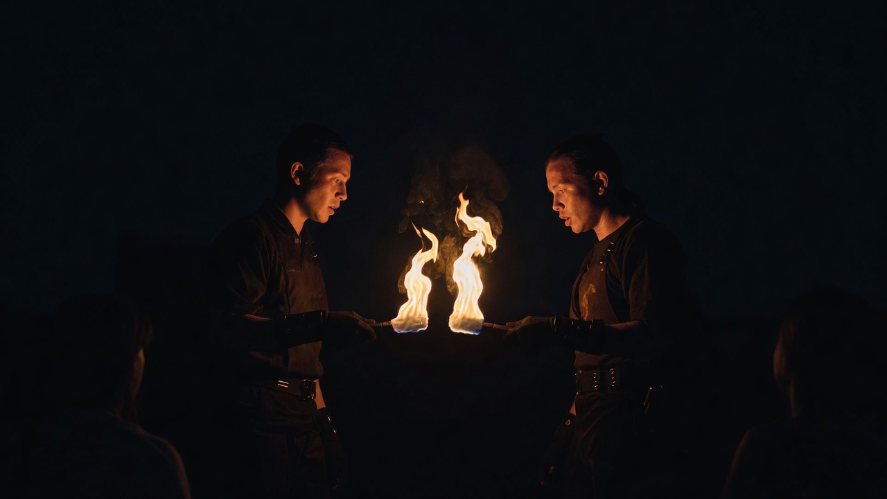 Two performers face each other in the dark, passing a lit fire staff, both faces softly lit by the shared flame.